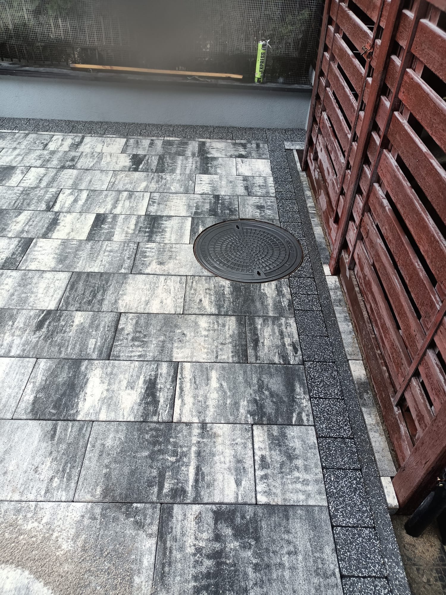 Ułożona, szara kostka brukowa z czarną obwódką i studzienką kanalizacyjną, sąsiadująca z drewnianym ogrodzeniem w odcieniach brązu. Widok z góry.