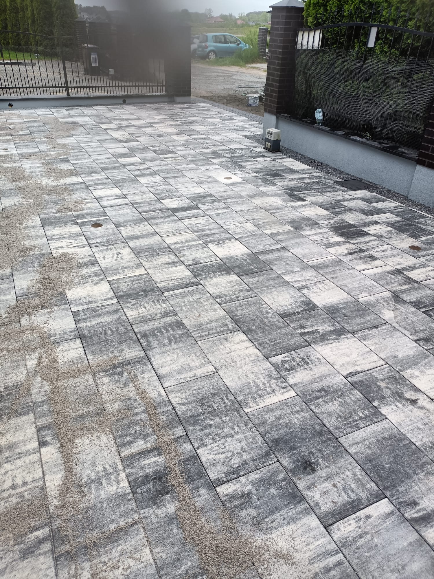 Nowoczesna, szara kostka brukowa ułożona na posesji, widoczne fragmenty ogrodzenia i bramy wjazdowej. Świeżo położona, posypana piaskiem.