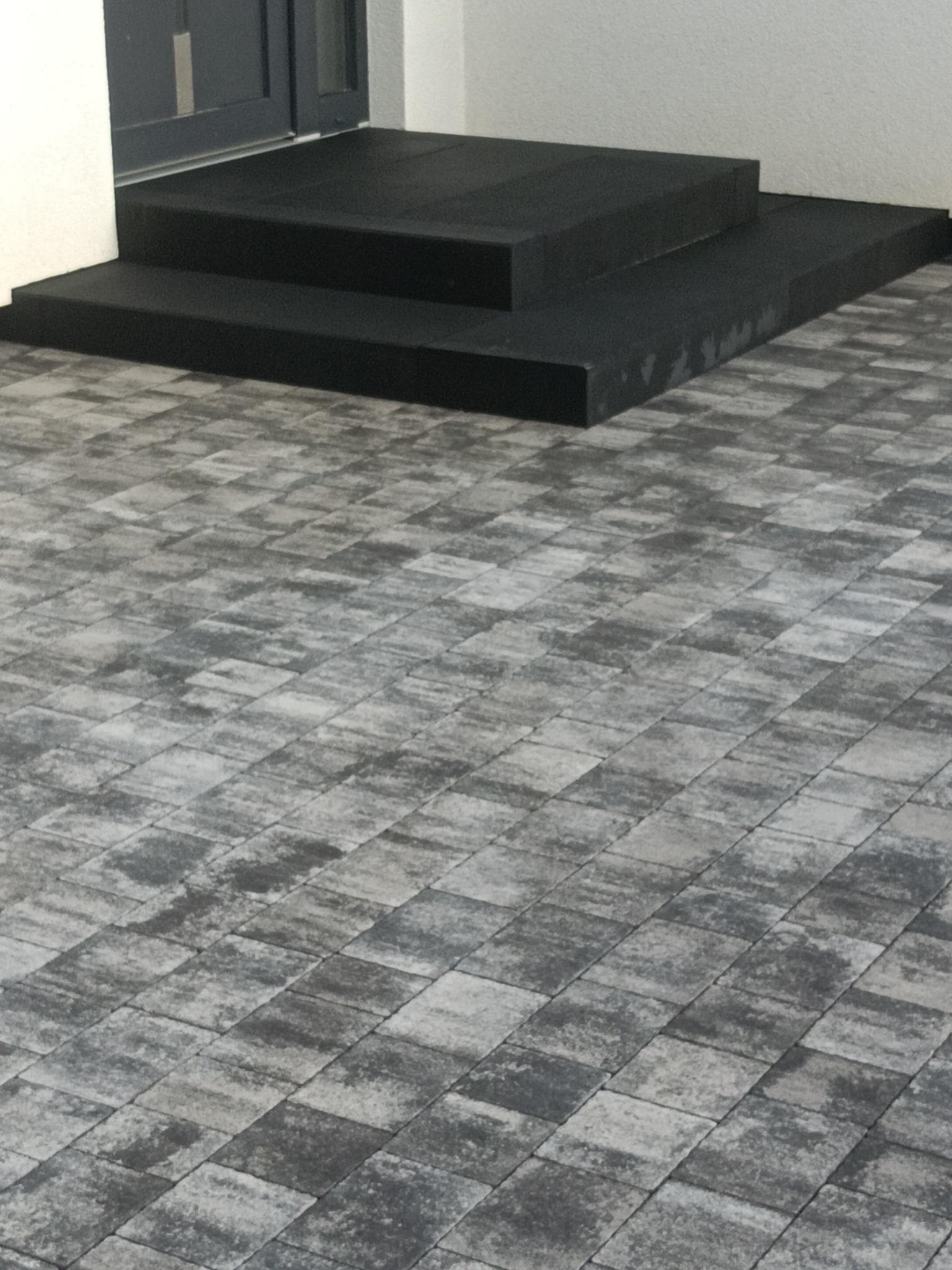Wykończone schody wejściowe z czarnej kostki brukowej, prowadzące do drzwi. Szara kostka brukowa na podłożu, ułożona w regularny wzór. Minimalistyczny design.