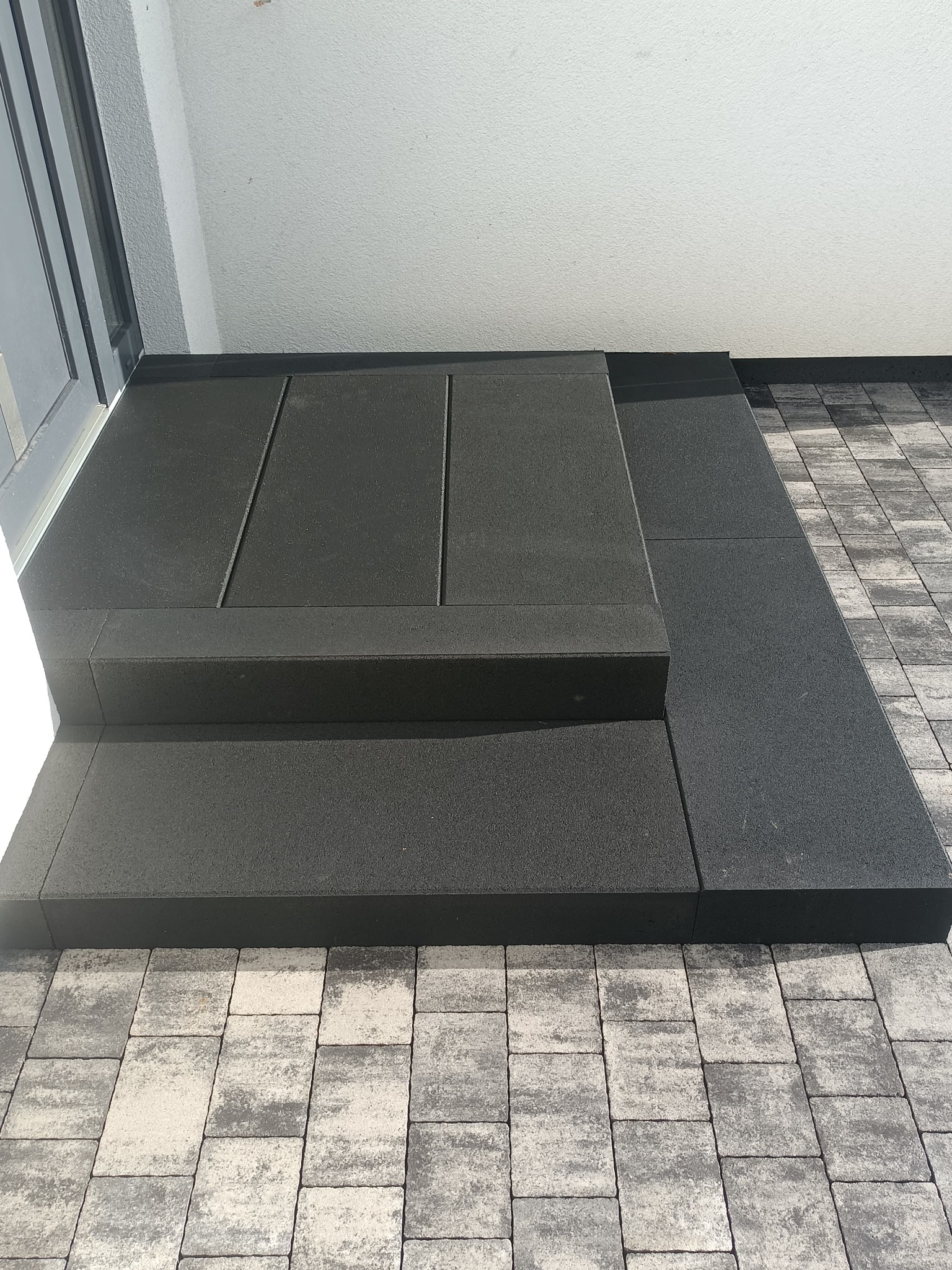 Schody z ciemnej kostki brukowej, z trzema stopniami, prowadzące na taras wyłożony jasnoszarą kostką. Widok z góry, podkreślający wzór ułożenia kostki.