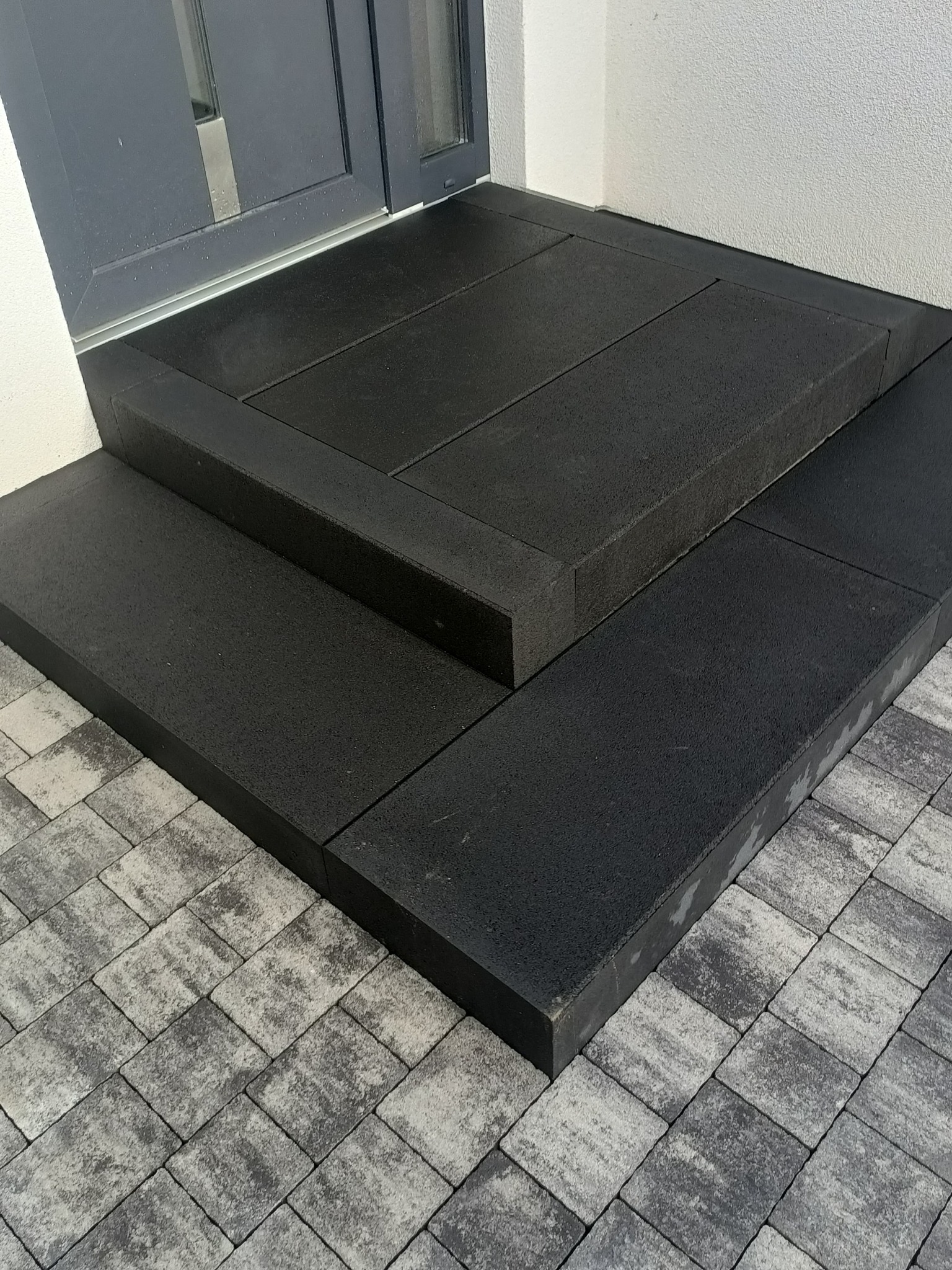 Nowoczesne, czarne stopnie wejściowe z kostki brukowej, prowadzące do szarych drzwi. Na dole widoczna szara kostka brukowa. Minimalistyczny design.