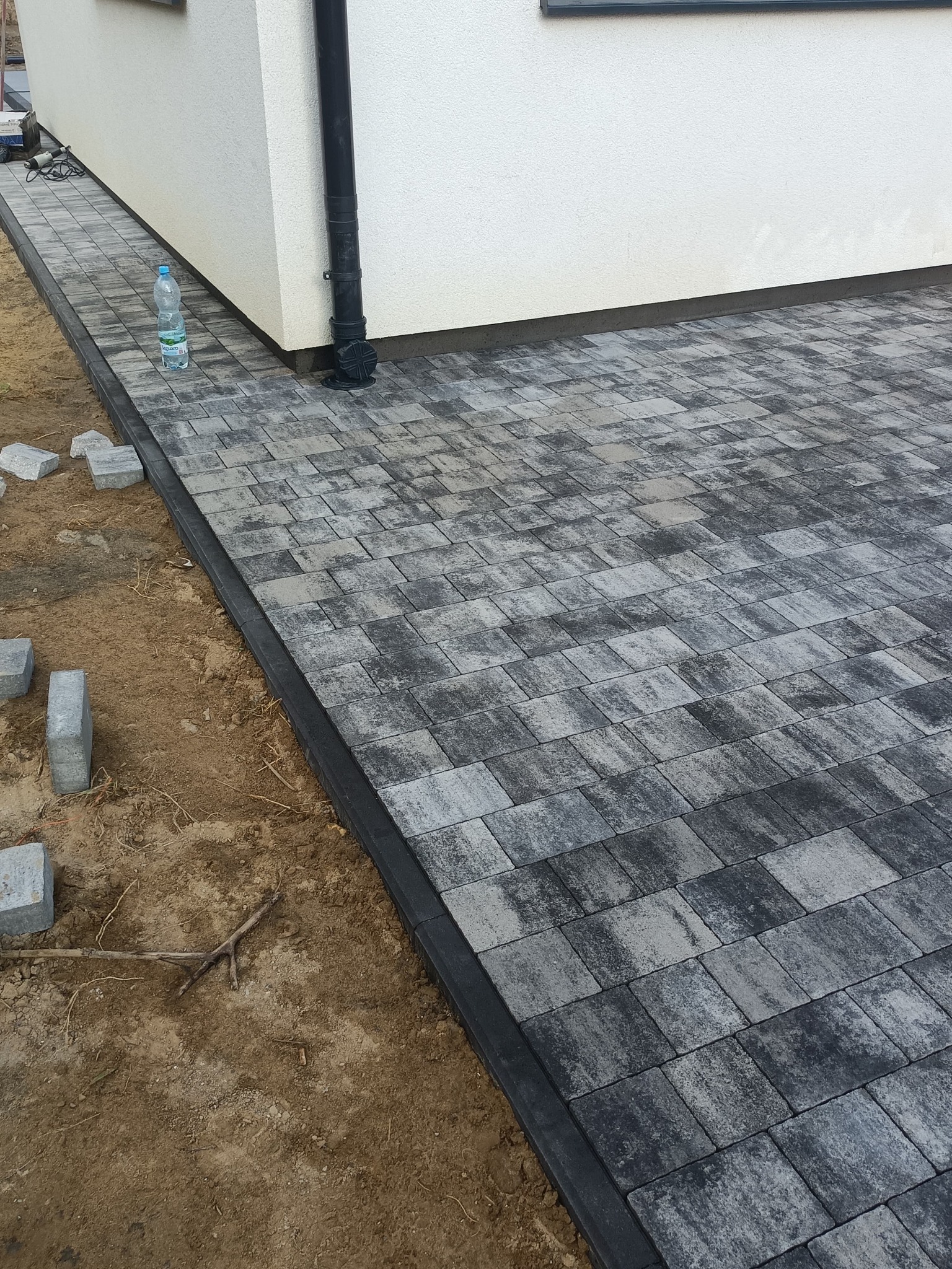 Ułożona kostka brukowa w odcieniach szarości przy białej ścianie budynku. Część kostki jeszcze nieułożona. Widoczna butelka wody.