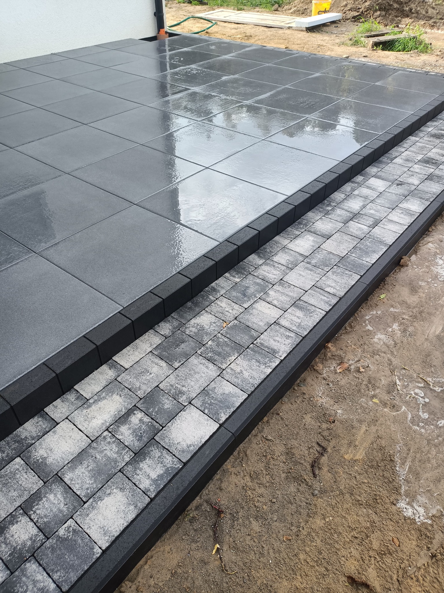 Szara, mokra nawierzchnia z dużych płyt i mniejszych kostek, obrzeża z czarnych elementów. Widoczne świeżo ułożone płyty i kostka brukowa.