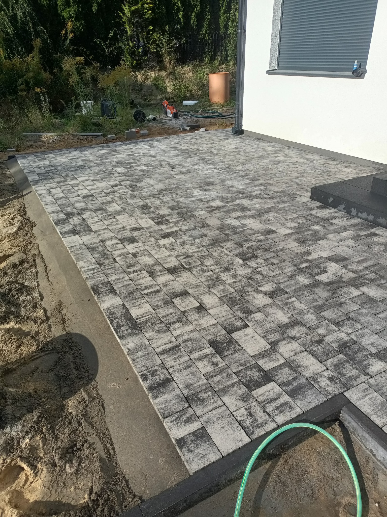 Ułożona kostka brukowa w odcieniach szarości na posesji, widoczna krawędź z obrzeżem, w tle trawnik i fragment domu. Użyto kostki o fakturze melanż.