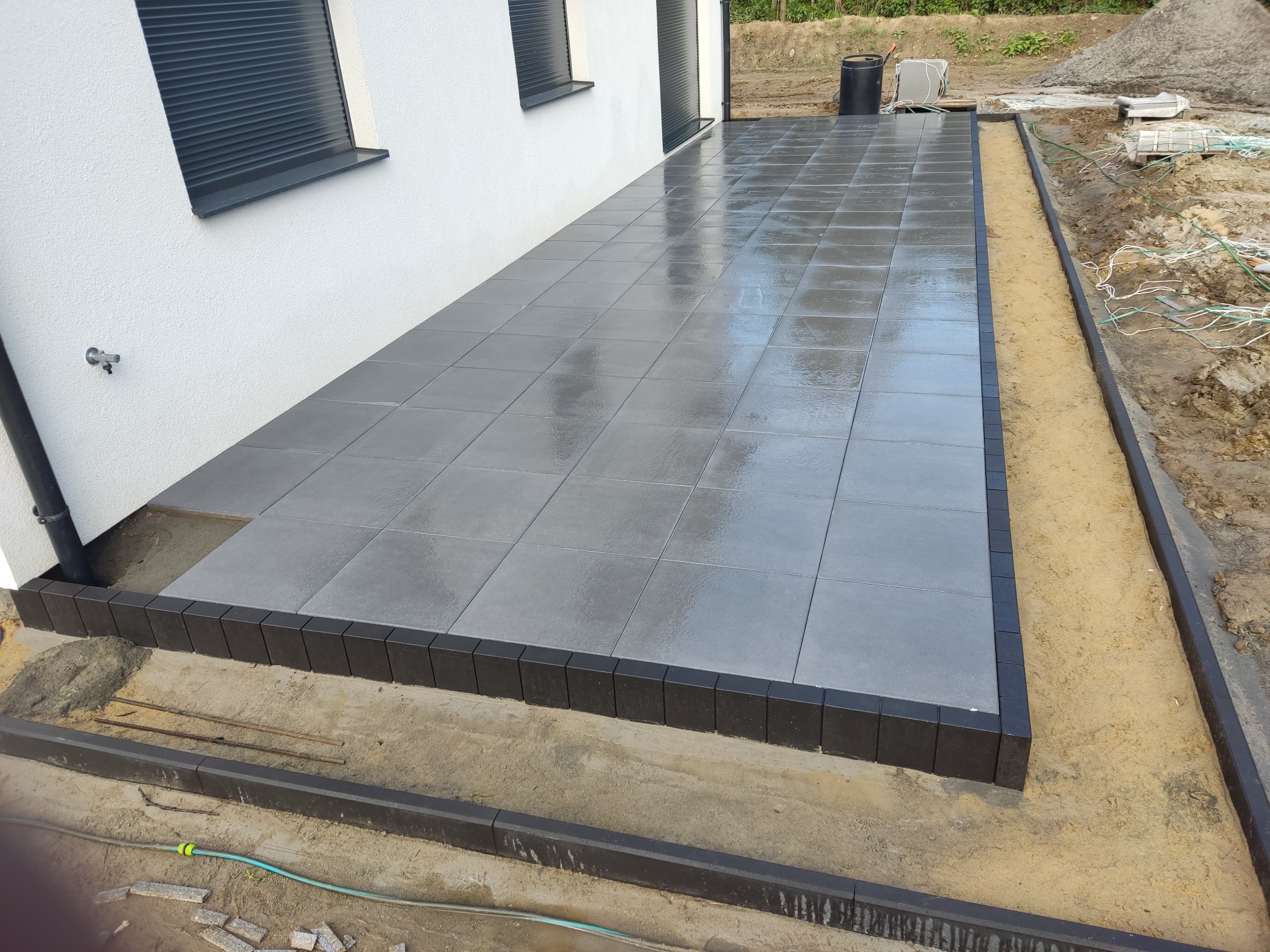 Nowoczesny taras z szarych płyt, obrzeża z ciemnej kostki. Widoczne przygotowania do dalszych prac ziemnych wokół tarasu. Świeżo ułożona nawierzchnia.