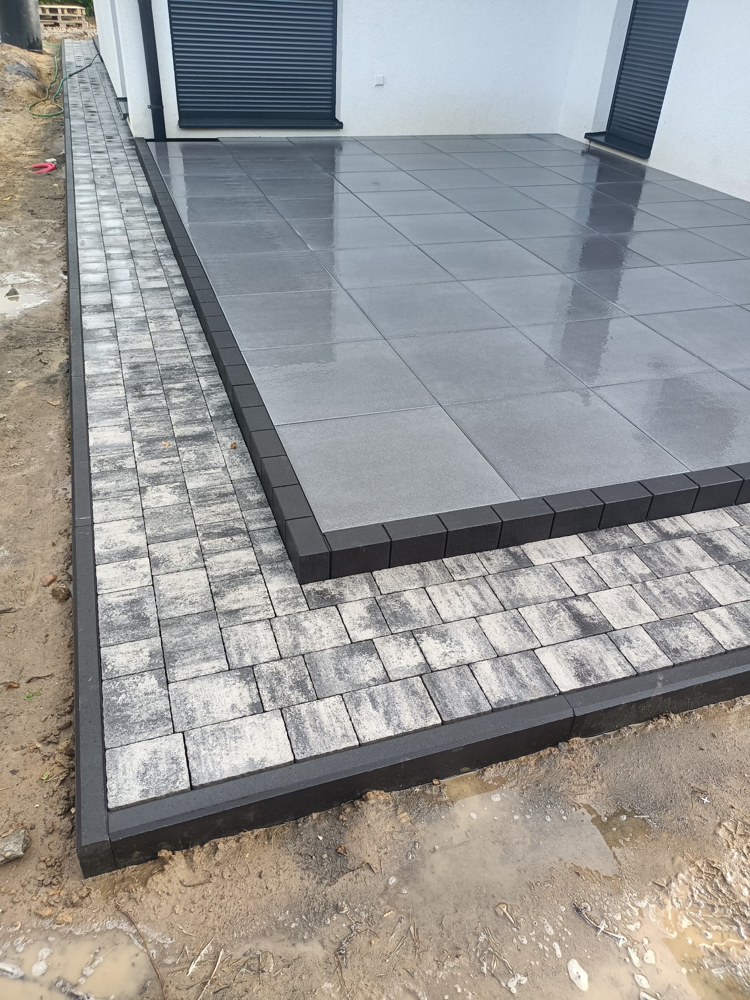 Nowoczesny taras z szarej kostki brukowej, obrzeża z ciemnego betonu. Widoczne świeże, mokre ślady po deszczu. Minimalistyczny design w otoczeniu domu.