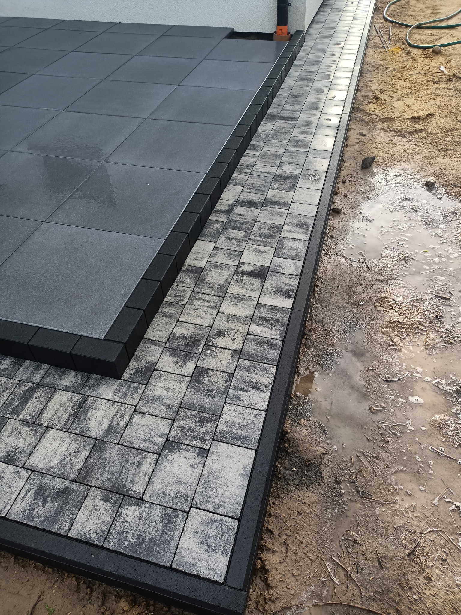 Ułożona, szara kostka brukowa z czarnym obrzeżem, przylegająca do ściany budynku i mokrego, piaszczystego gruntu. Widoczne różne faktury kostki.