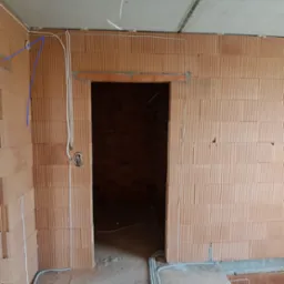 Wewnętrzne okablowanie elektryczne w surowym stanie, widoczne przewody prowadzone po ceglanej ścianie i pod sufitem w nowo budowanym pomieszczeniu.