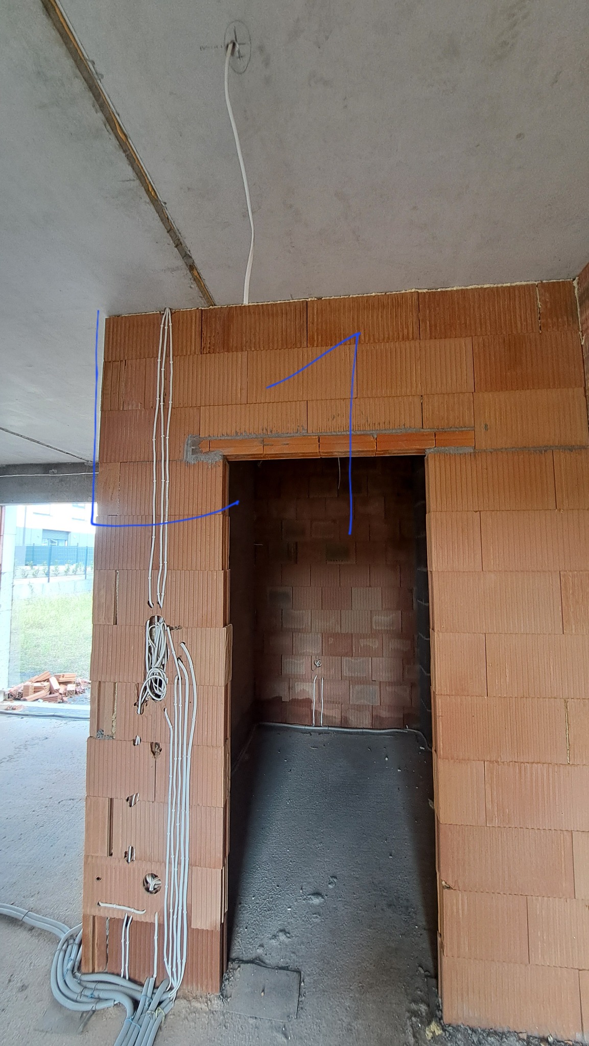 Surowy stan: przewody elektryczne prowadzone wzdłuż ściany z cegły dziurawki wewnątrz budynku w trakcie budowy, widoczne otwory montażowe i szare rury kablowe na podłodze.
