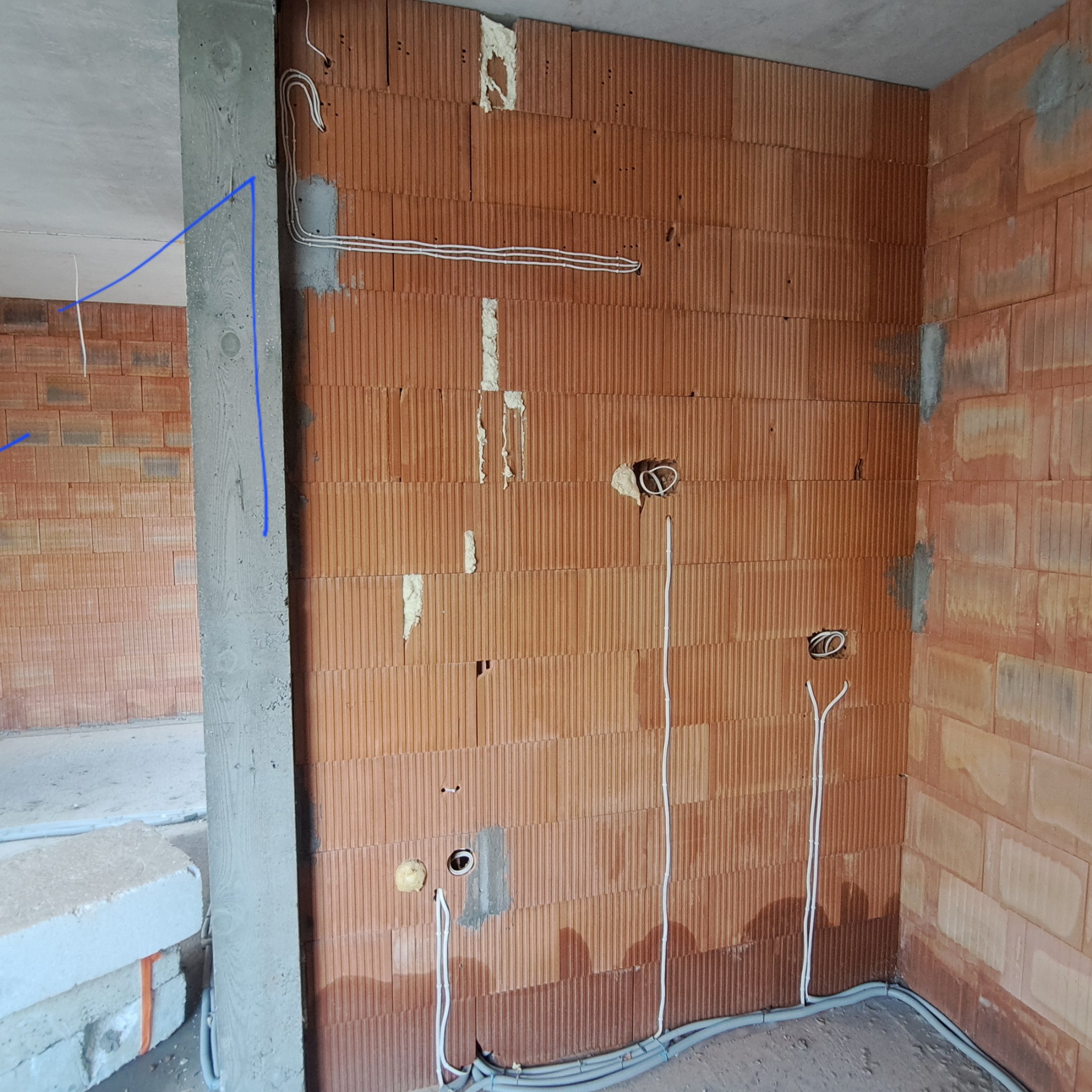 Surowy mur z cegły dziurawki z widocznymi przewodami elektrycznymi wychodzącymi ze ściany, częściowo zabezpieczonymi pianką montażową, oraz szarymi rurkami przy podłodze, w tle widoczny betonowy...