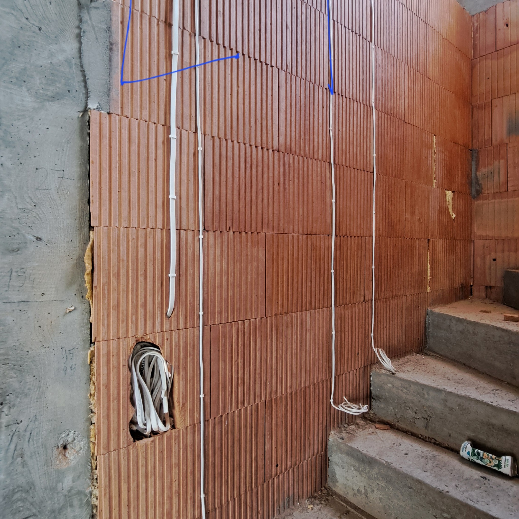 Ściana z czerwonej cegły z pionowo poprowadzonymi białymi kablami elektrycznymi, niebieskie linie zaznaczone na ścianie, otwór w ścianie z widocznymi kablami, fragment betonowych schodów.
