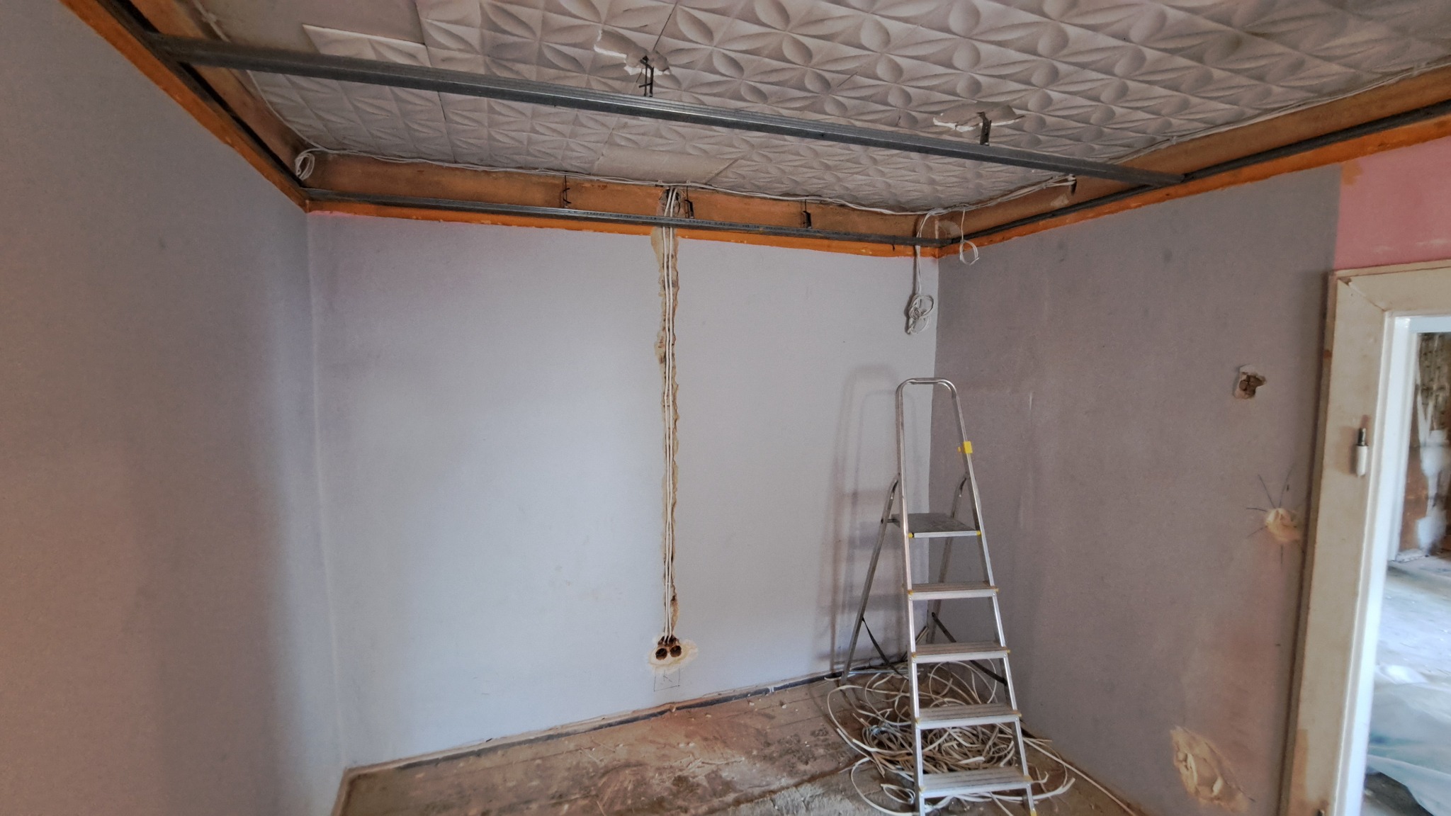 Wnętrze pokoju w trakcie remontu z widocznymi przewodami elektrycznymi wystającymi ze ściany, metalową drabinką i częściowo zamontowanym sufitem podwieszanym z dekoracyjnymi panelami.