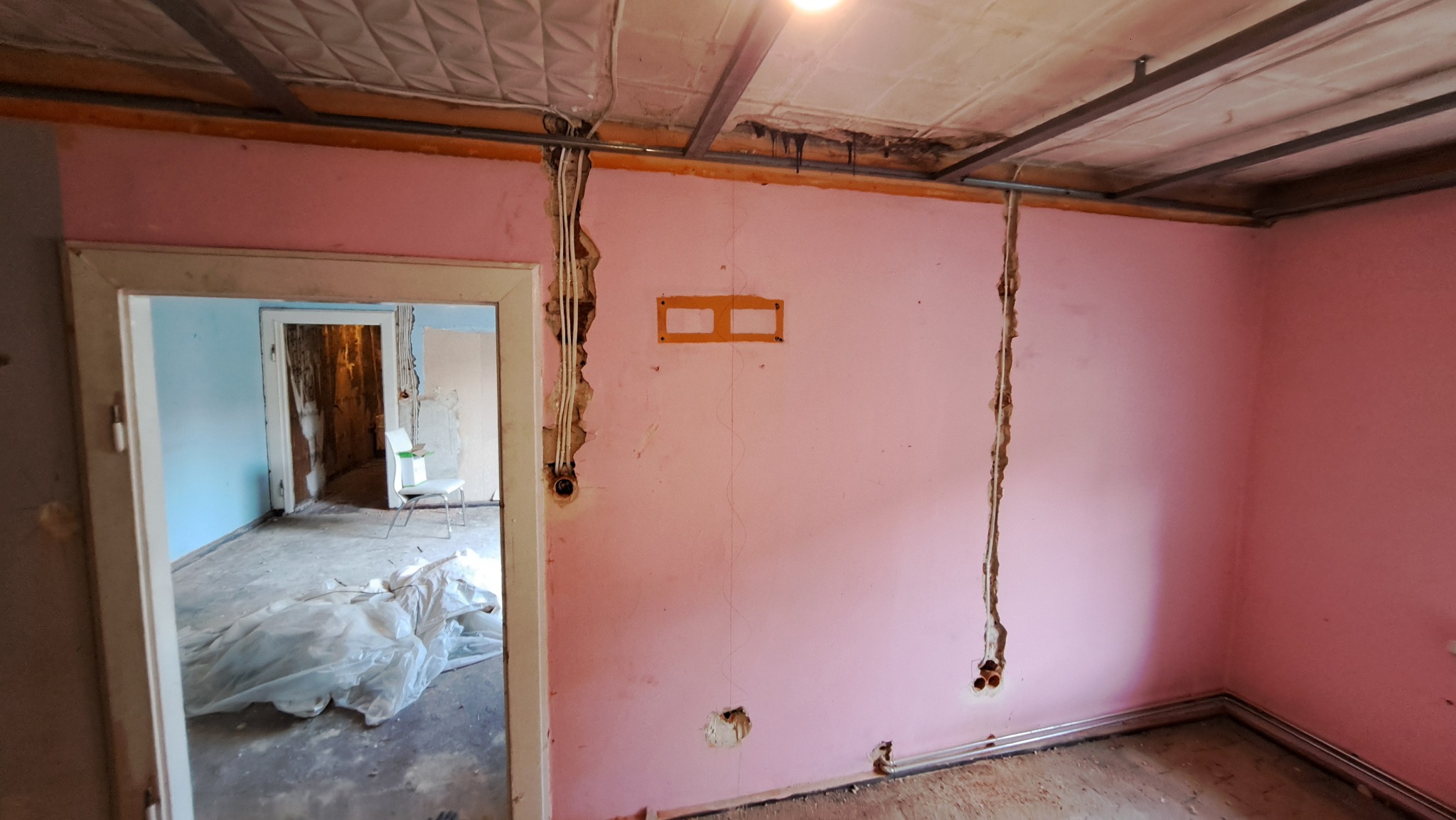 Różowy pokój w trakcie remontu z widocznymi bruzdami w ścianie przygotowanymi pod przewody elektryczne, odsłonięta instalacja elektryczna w ścianie, fragment podwieszanego sufitu z widocznymi...