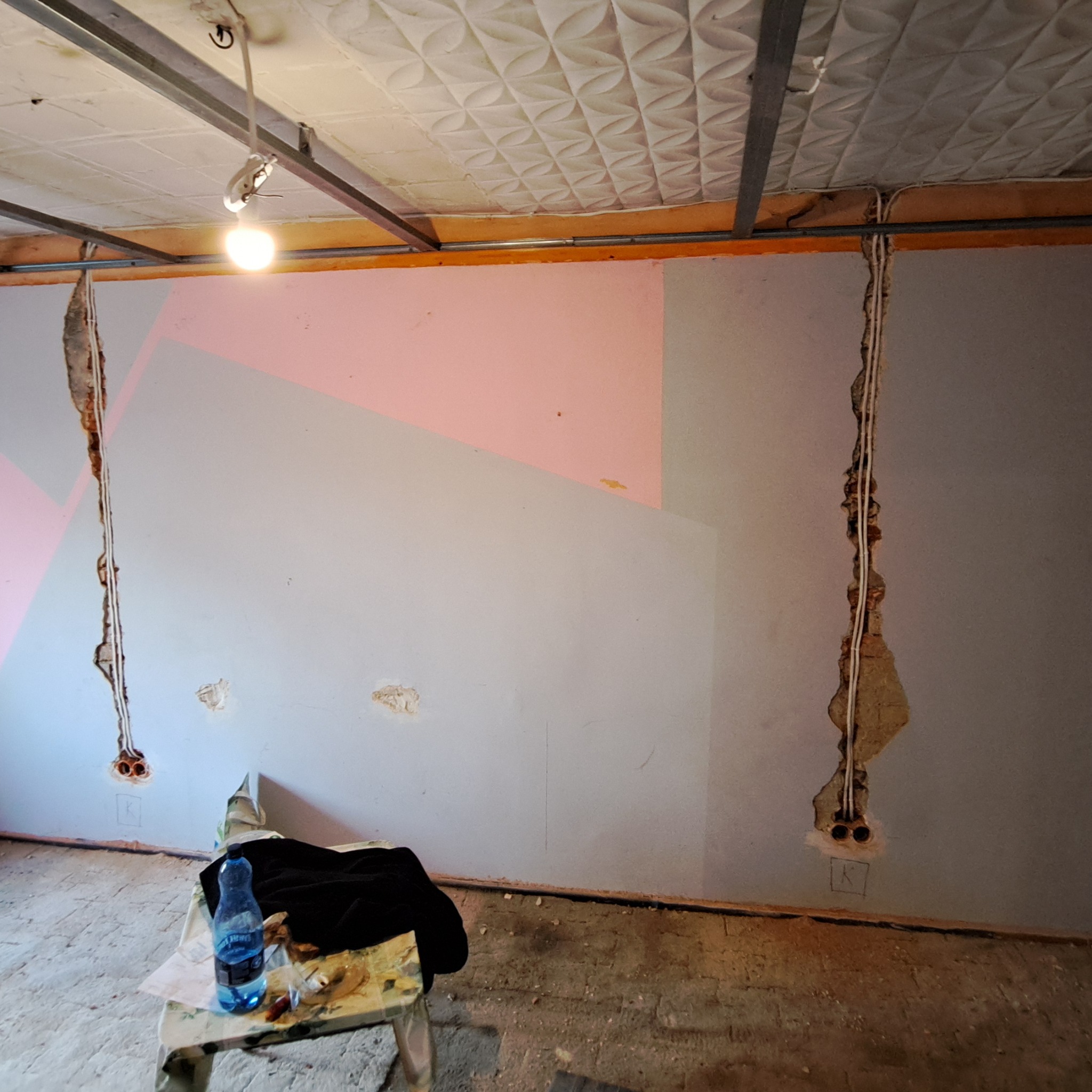 Wnętrze w trakcie remontu; widoczne kable elektryczne wystające ze ścian, ściany z geometrycznym wzorem w odcieniach szarości i różu, oraz sufit podwieszany z metalową konstrukcją i oświetleniem...