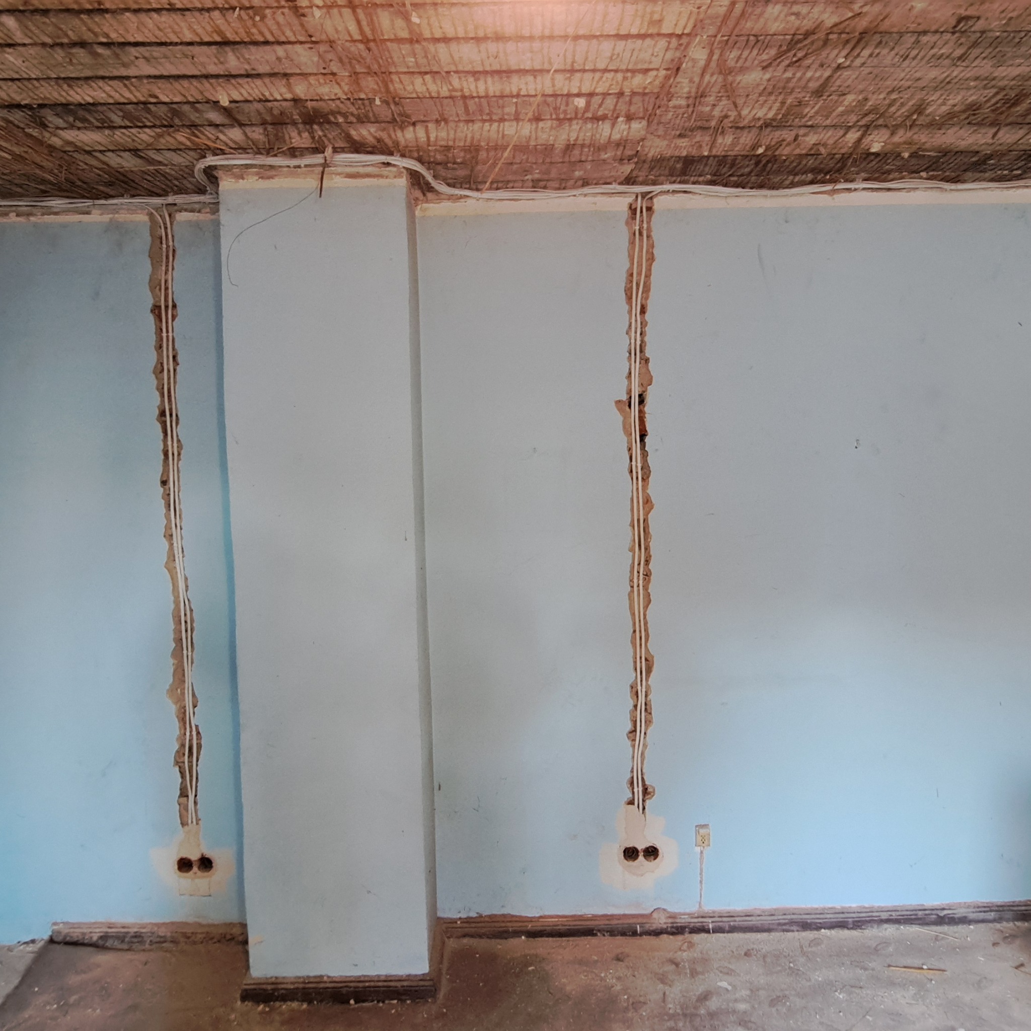 Ściana w trakcie bruzdowania pod przewody elektryczne, widoczne puste puszki pod gniazdka, wystające przewody oraz surowy strop z desek.