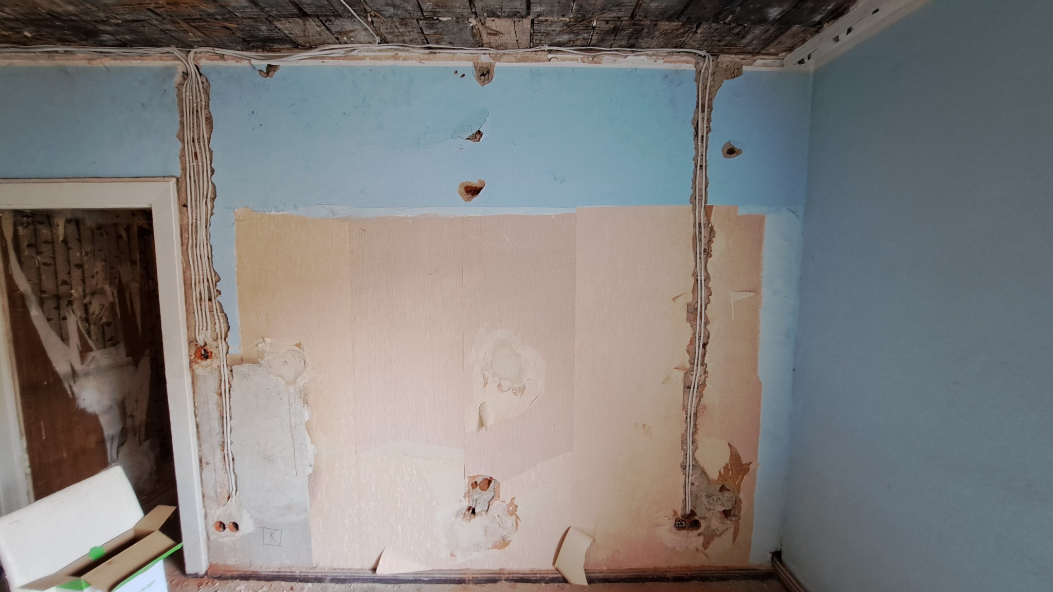 Ściana w trakcie remontu z przewodami elektrycznymi w bruzdach, widoczne stare warstwy farby i tapety, odsłonięta instalacja elektryczna przed położeniem tynku.