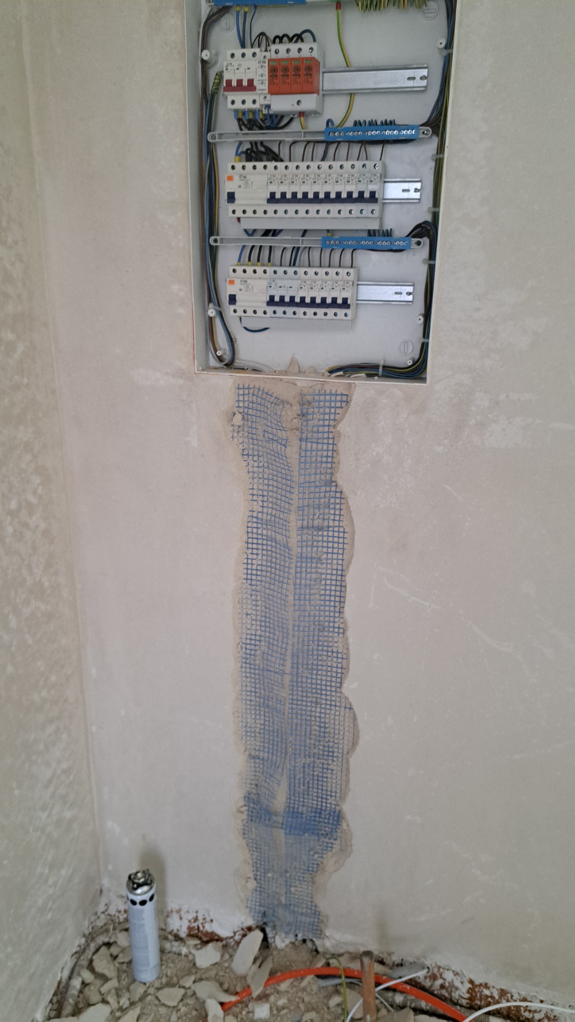 Otwarta skrzynka elektryczna z zabezpieczeniami i przewodami, widoczna siatka wzmacniająca na ścianie, przygotowana do tynkowania, oraz gruz na podłodze.