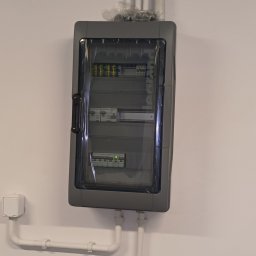 GRU Innovations PRZEMYSŁAW WOJTYŁA - Szara skrzynka elektryczna Legrand z przezroczystymi drzwiczkami, zamontowana na białej ścianie, z widocznymi przewodami i rurkami instalacyjnymi.