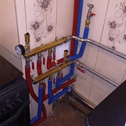 GRU Innovations PRZEMYSŁAW WOJTYŁA - Rozdzielacz instalacji grzewczej z manometrem i zaworami, rury w kolorze czerwonym i niebieskim, widoczne łączenia i fragment pieca w pomieszczeniu z ceramiczną okładziną ścienną.