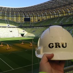 GRU Innovations PRZEMYSŁAW WOJTYŁA - Biały kask budowlany z napisem 'GRU' na tle zielonego stadionu z pustymi trybunami i oświetleniem boiska
