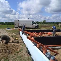GRU Innovations PRZEMYSŁAW WOJTYŁA - Prace ziemne i przygotowanie do instalacji kanalizacyjnej przy fundamentach domu jednorodzinnego, widoczne rury kanalizacyjne, izolacja fundamentów i pracujący hydraulicy.