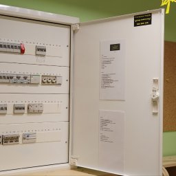 GRU Innovations PRZEMYSŁAW WOJTYŁA - Otwarta skrzynka elektryczna z bezpiecznikami i wyłącznikami, zamontowana na ścianie w pomieszczeniu o zielonych ścianach, z naklejką firmy elektrycznej na drzwiach.