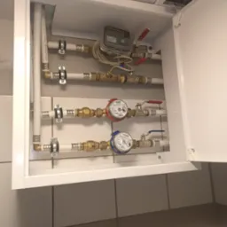 Wewnętrzny widok szafki z instalacją hydrauliczną w Barniewicach, widoczne rury, zawory, liczniki wody i ciepłomierz elektroniczny. Szafka z otwartymi drzwiczkami, na ścianie płytki ceramiczne.