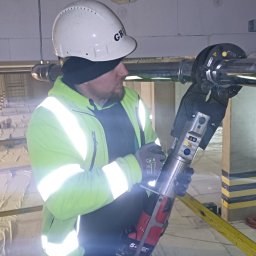 GRU Innovations PRZEMYSŁAW WOJTYŁA - Pracownik w kasku i odblaskowej kurtce zaciska rurę hydrauliczną za pomocą specjalistycznej prasy akumulatorowej w surowym wnętrzu budynku z widocznymi otworami w ścianie.