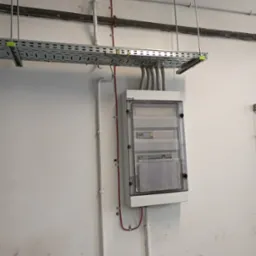 Szara szafa elektryczna z przezroczystymi drzwiami, zamontowana na białej ścianie, z widocznymi przewodami w metalowej korycie podwieszonym pod sufitem i rurami instalacyjnymi.