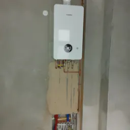 Biały piec gazowy Junkers zamontowany na surowej, szarej ścianie z widocznymi miedzianymi rurami i fragmentem instalacji podłogowej w ścianie.