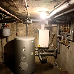 GRU Innovations PRZEMYSŁAW WOJTYŁA - Instalacja kotła gazowego w piwnicy, widoczne rury, zawory, zbiornik buforowy, ściany z surowego betonu i przewody elektryczne.