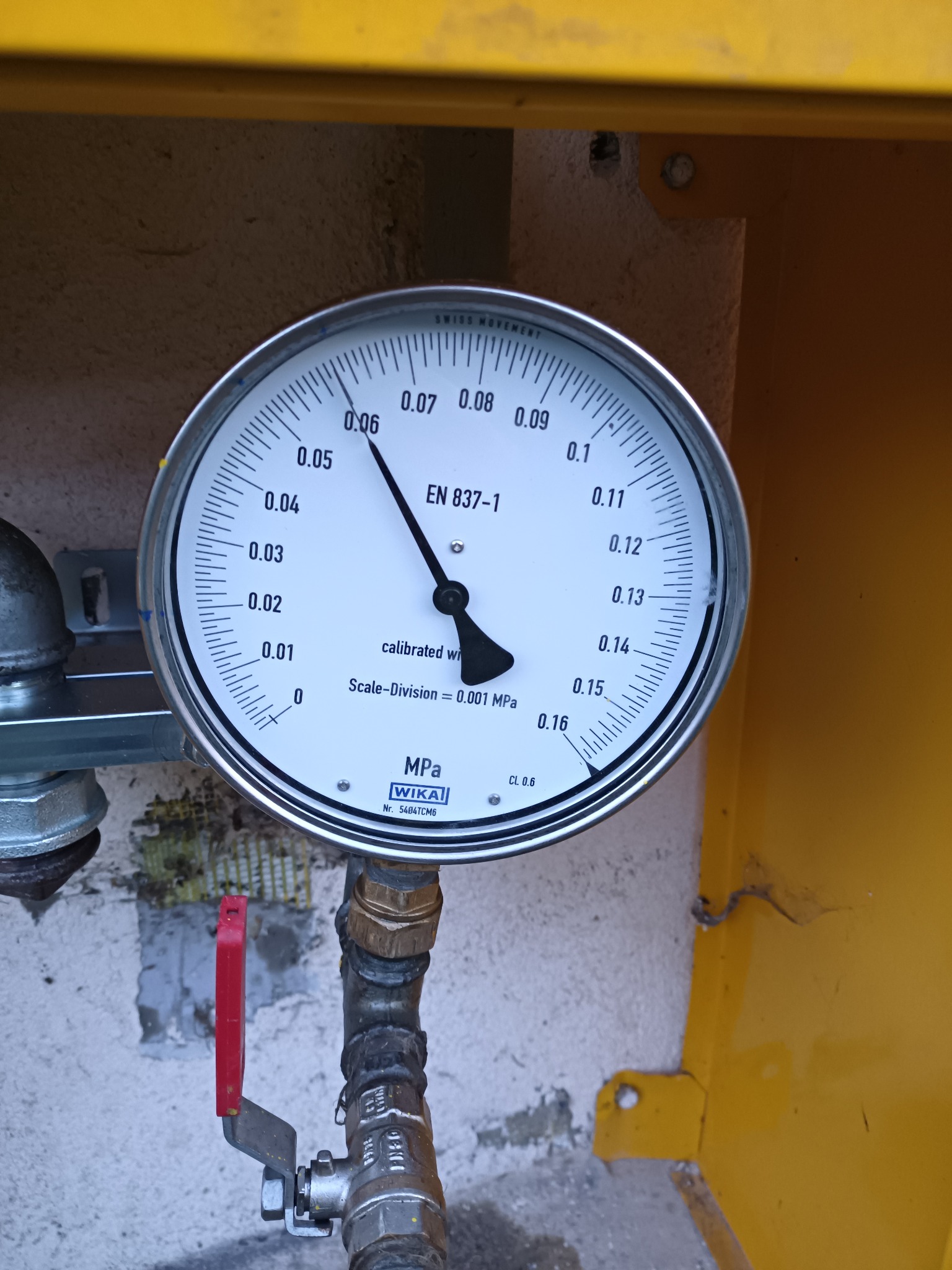 Manometr WIKA wskazujący ciśnienie w instalacji gazowej, zamontowany na żółtej obudowie, z widocznym zaworem odcinającym z czerwoną rączką.