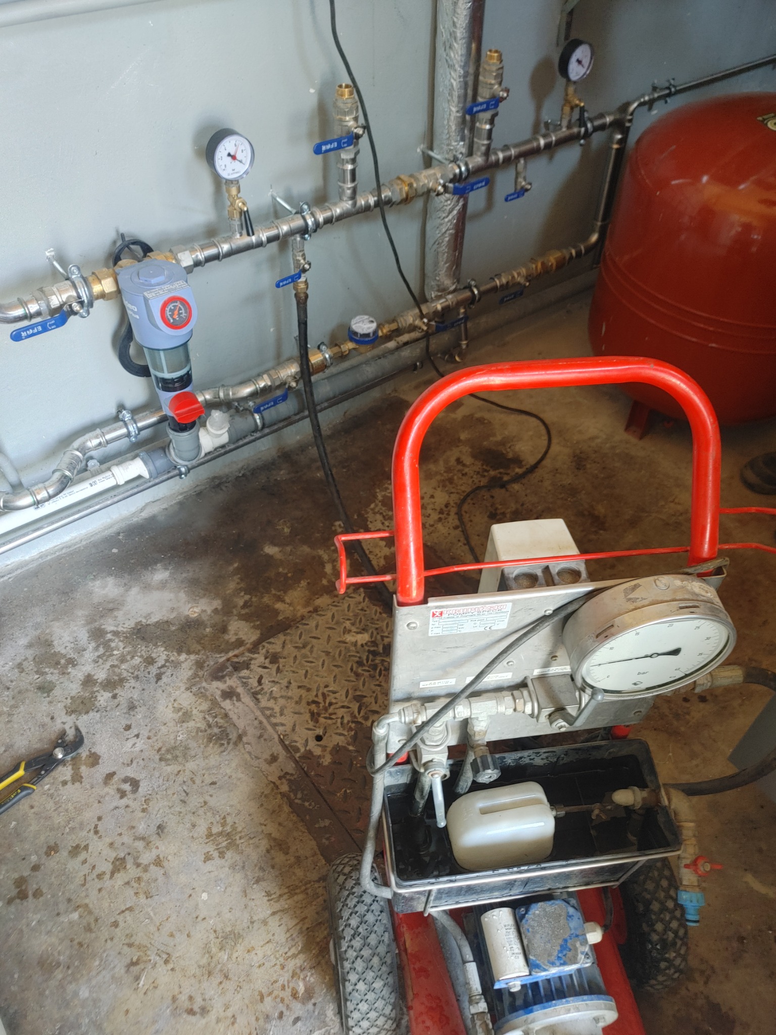 Próba ciśnieniowa instalacji hydraulicznej: widoczny manometr na urządzeniu do testowania szczelności, rury z zaworami i manometrami zamontowane na ścianie, zbiornik wyrównawczy w tle.