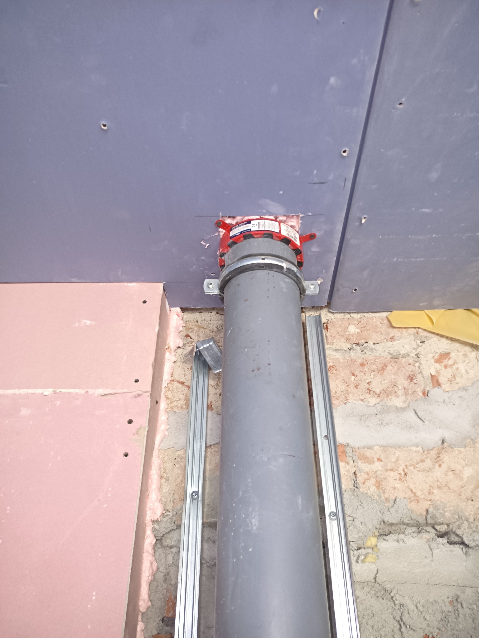 Szara rura kanalizacyjna z czerwoną obejmą, zamocowana do sufitu z płyt gipsowo-kartonowych, widoczne metalowe profile i fragment muru z cegły.