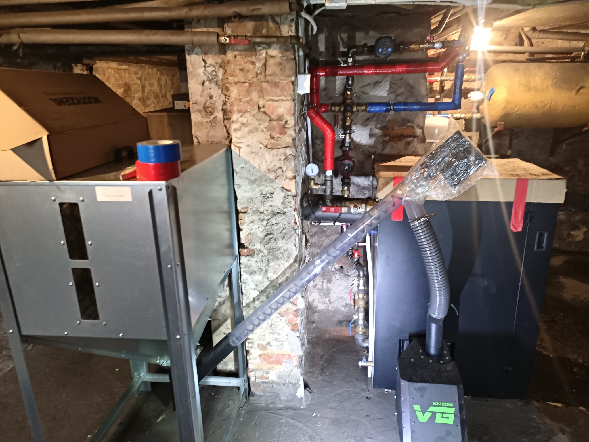 Nowoczesny piec na paliwo stałe z podajnikiem ślimakowym w kotłowni, widoczne rury instalacji grzewczej i fragment ściany z cegły.