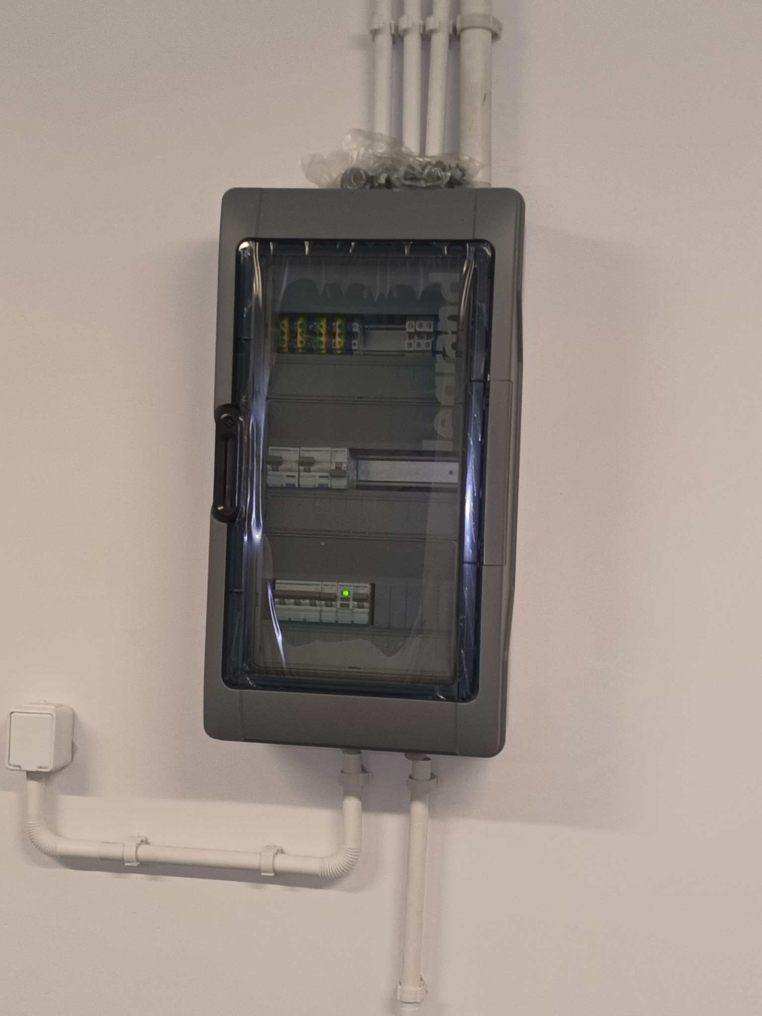 Szara skrzynka elektryczna Legrand z przezroczystymi drzwiczkami, zamontowana na białej ścianie, z widocznymi przewodami i rurkami instalacyjnymi.