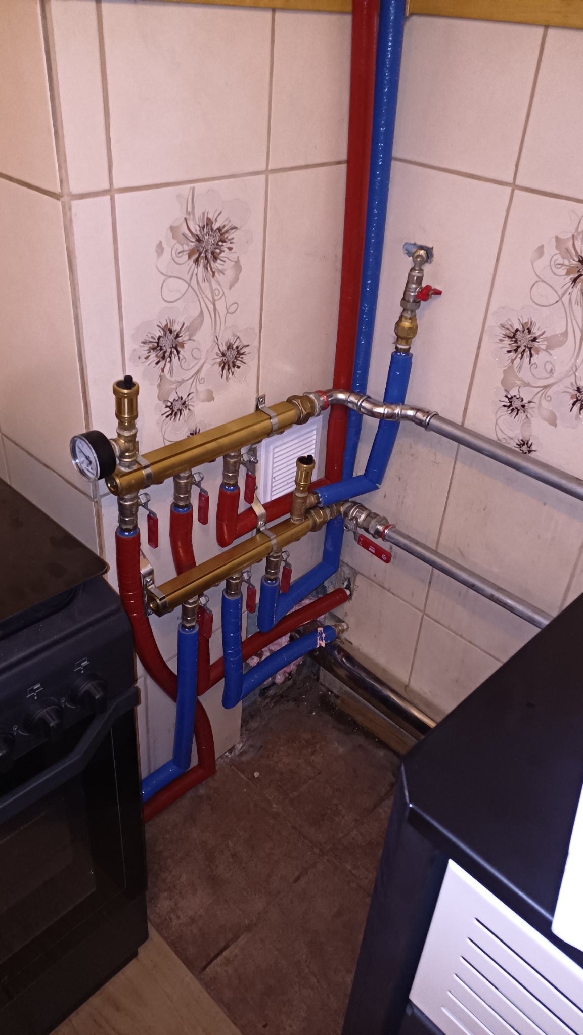 Rozdzielacz instalacji grzewczej z manometrem i zaworami, rury w kolorze czerwonym i niebieskim, widoczne łączenia i fragment pieca w pomieszczeniu z ceramiczną okładziną ścienną.