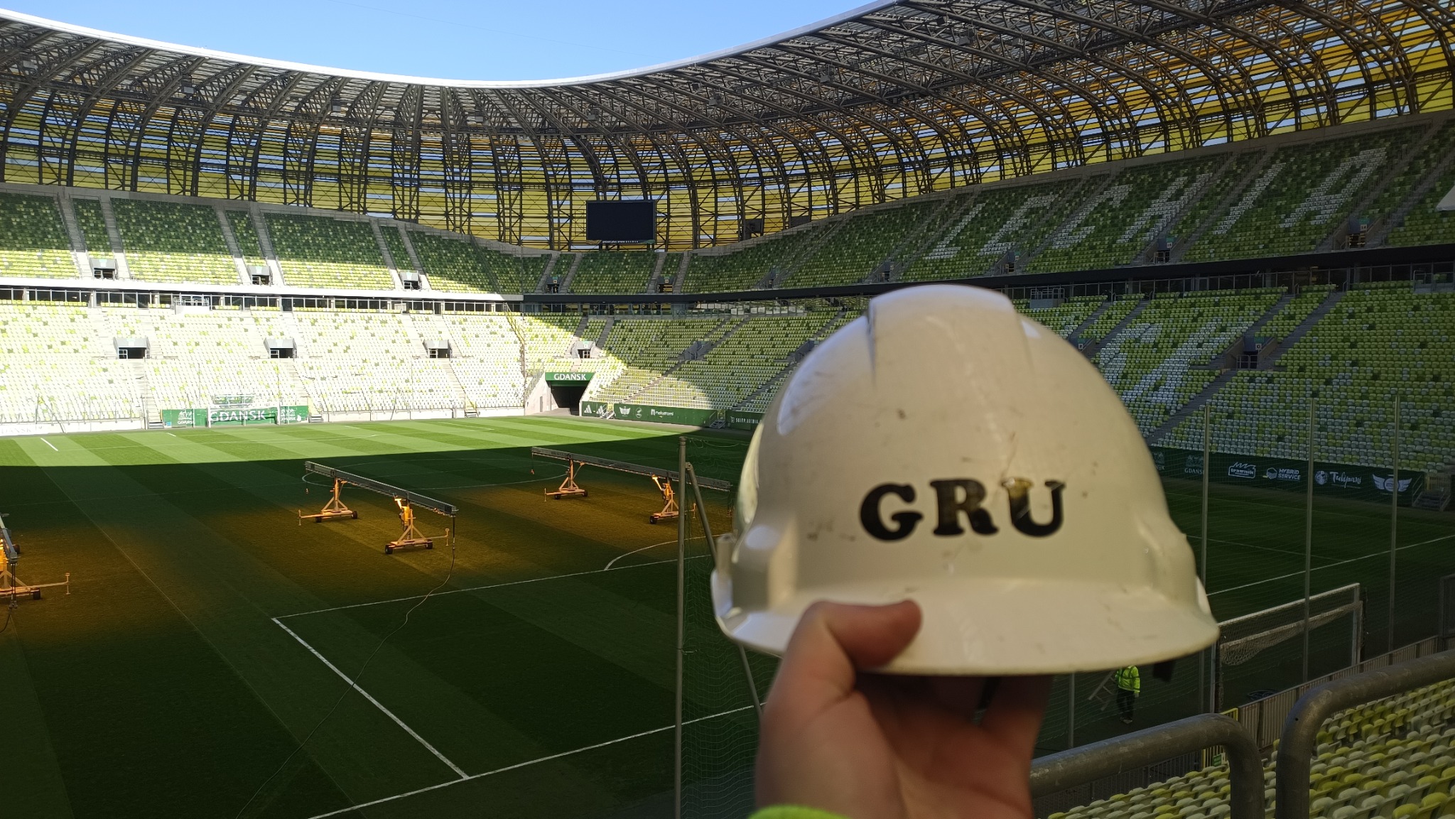 Biały kask budowlany z napisem 'GRU' na tle zielonego stadionu z pustymi trybunami i oświetleniem boiska