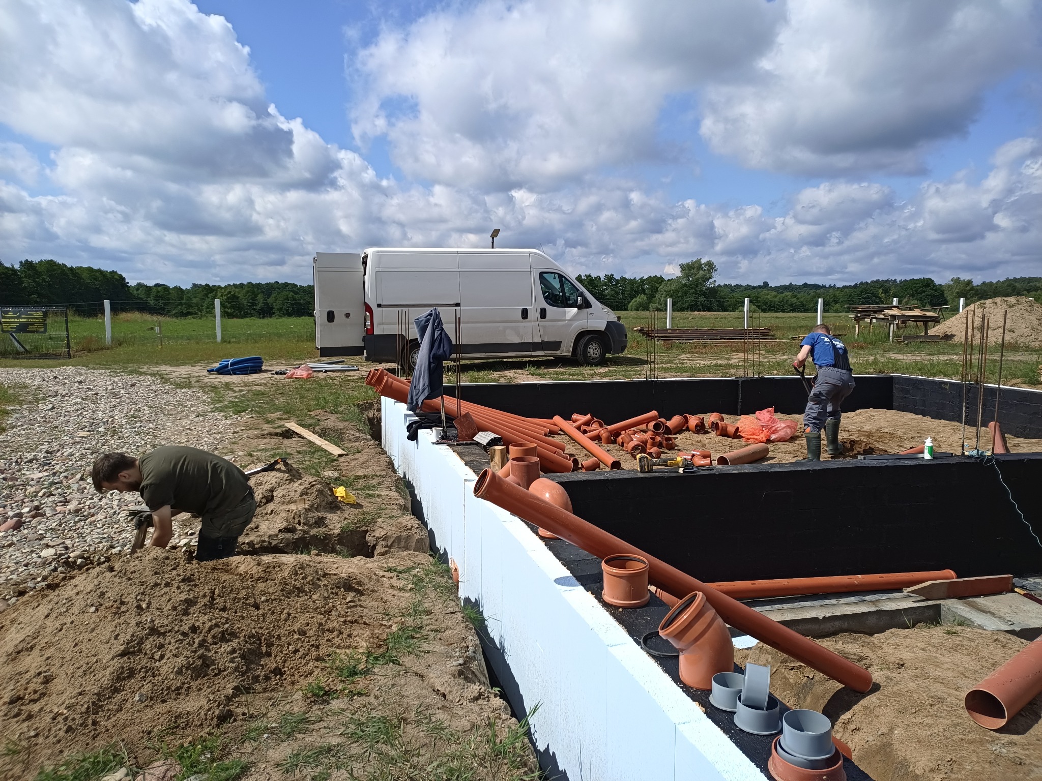Prace ziemne i przygotowanie do instalacji kanalizacyjnej przy fundamentach domu jednorodzinnego, widoczne rury kanalizacyjne, izolacja fundamentów i pracujący hydraulicy.