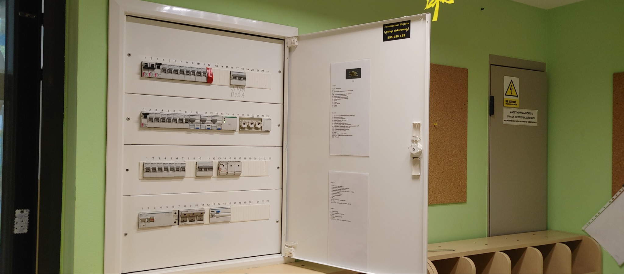 Otwarta skrzynka elektryczna z bezpiecznikami i wyłącznikami, zamontowana na ścianie w pomieszczeniu o zielonych ścianach, z naklejką firmy elektrycznej na drzwiach.