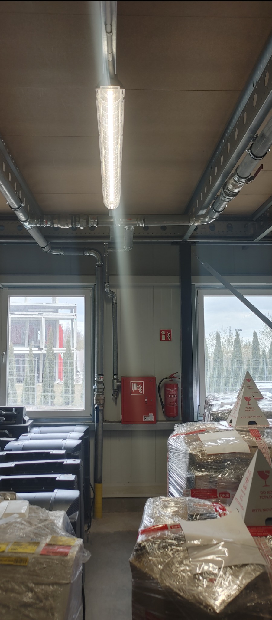 Wnętrze magazynu z instalacją rur, oświetleniem LED i zapakowanymi towarami, widok na okno z drzewami.
