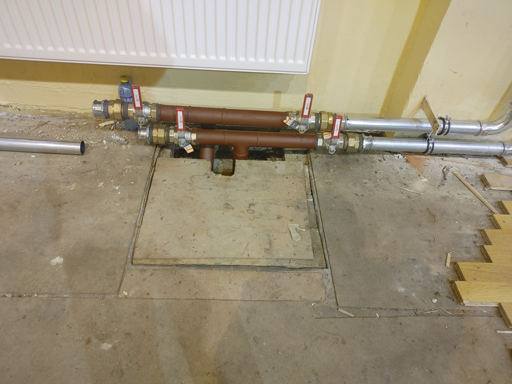 Podłączenie rur grzewczych z zaworami, widoczne na tle podłogi w trakcie remontu, z odsłoniętym włazem rewizyjnym i fragmentami parkietu.