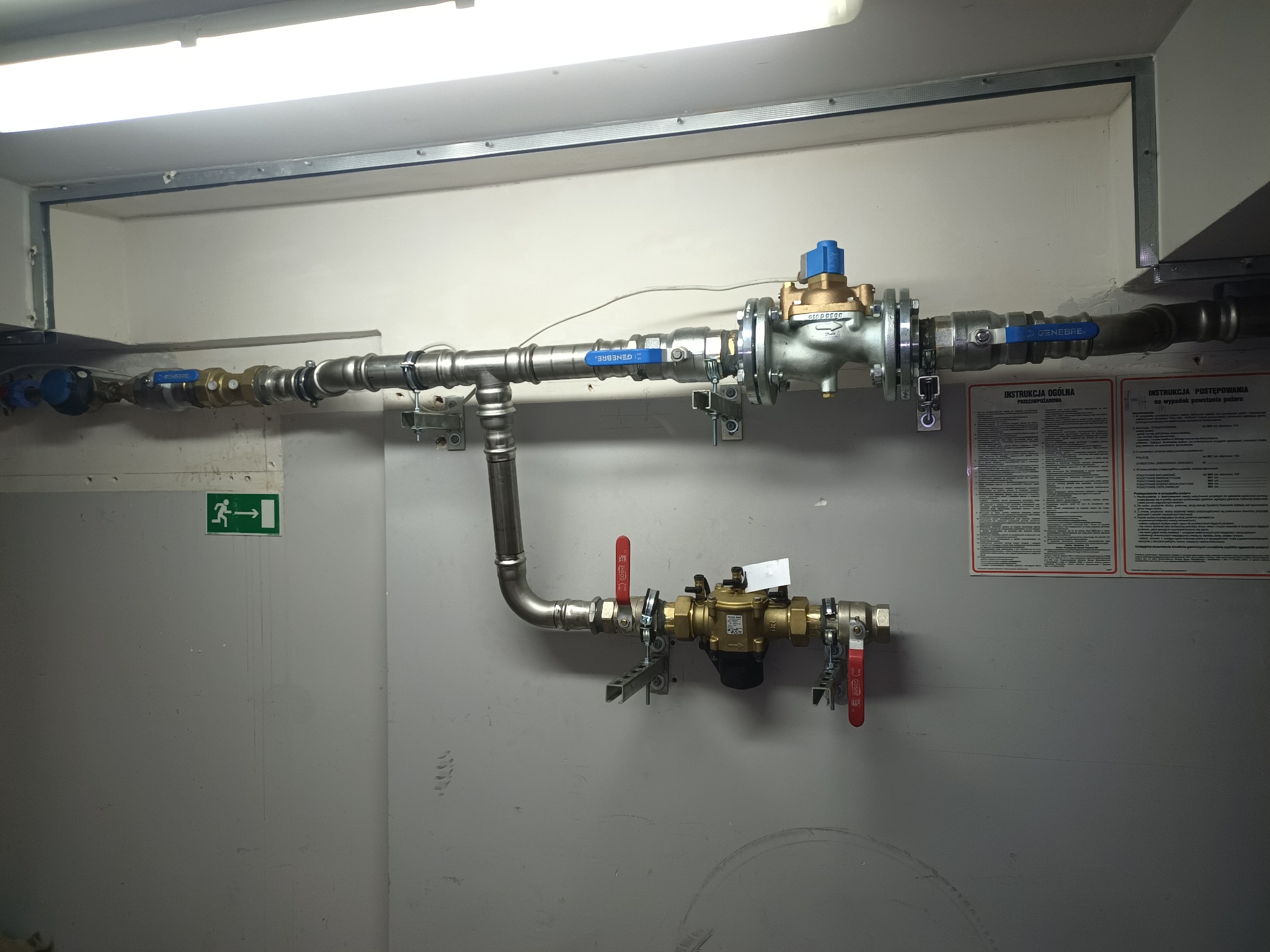 Zestaw zaworów i rur, w tym reduktor ciśnienia, zamontowany na szarej ścianie, z widoczną instrukcją obsługi i znakiem ewakuacyjnym.