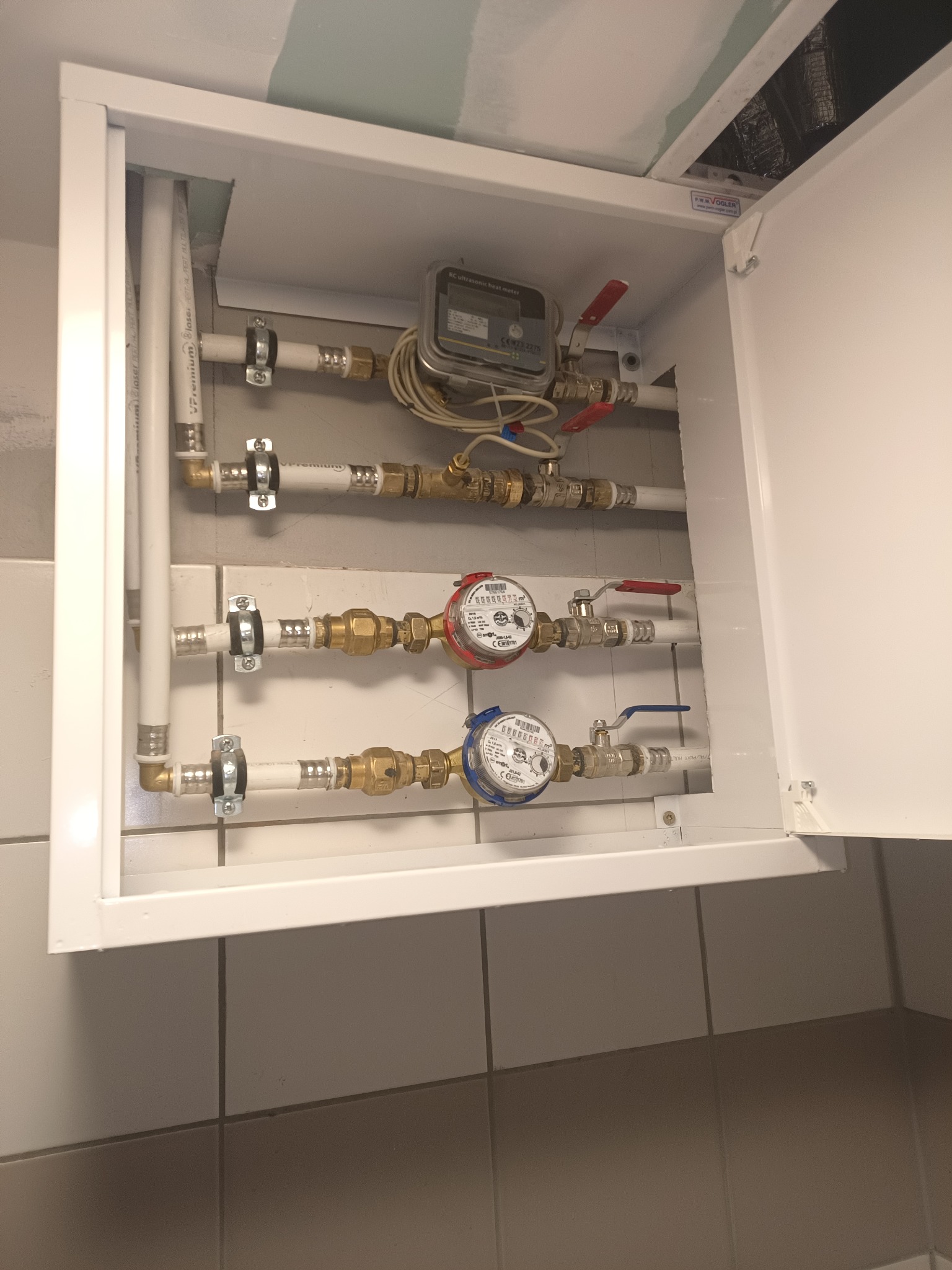 Wewnętrzny widok szafki z instalacją hydrauliczną w Barniewicach, widoczne rury, zawory, liczniki wody i ciepłomierz elektroniczny. Szafka z otwartymi drzwiczkami, na ścianie płytki ceramiczne.