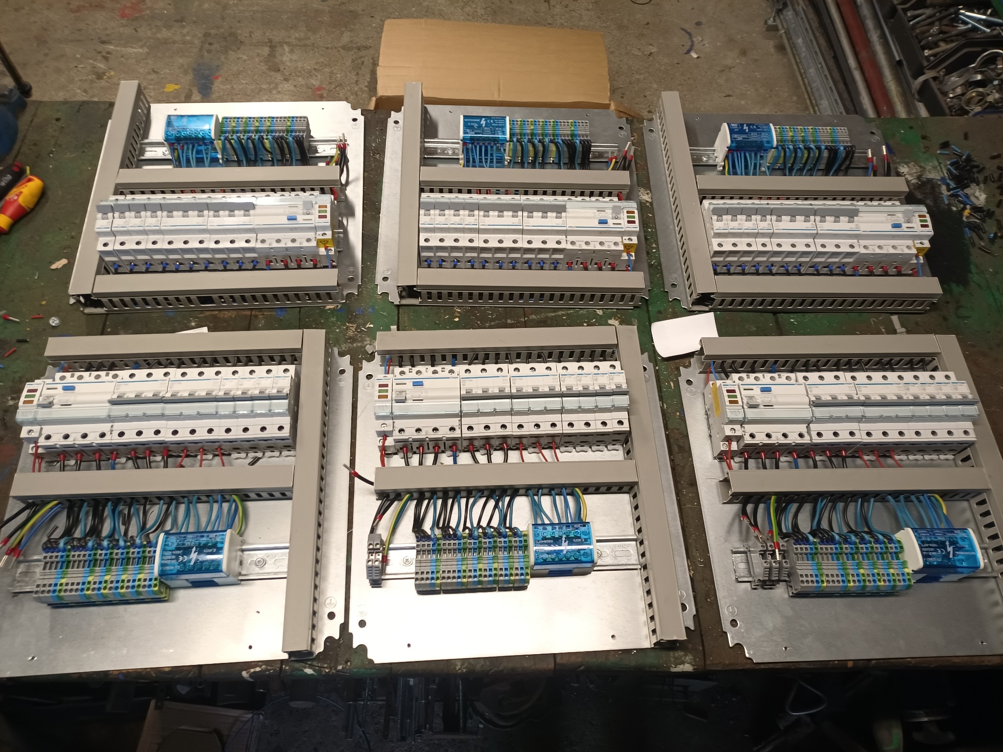 Sześć rozdzielnic elektrycznych w szarych obudowach, z widocznymi bezpiecznikami, listwami zaciskowymi i przewodami o różnych kolorach, ułożonych na roboczym stole z widocznymi śladami użytkowania.