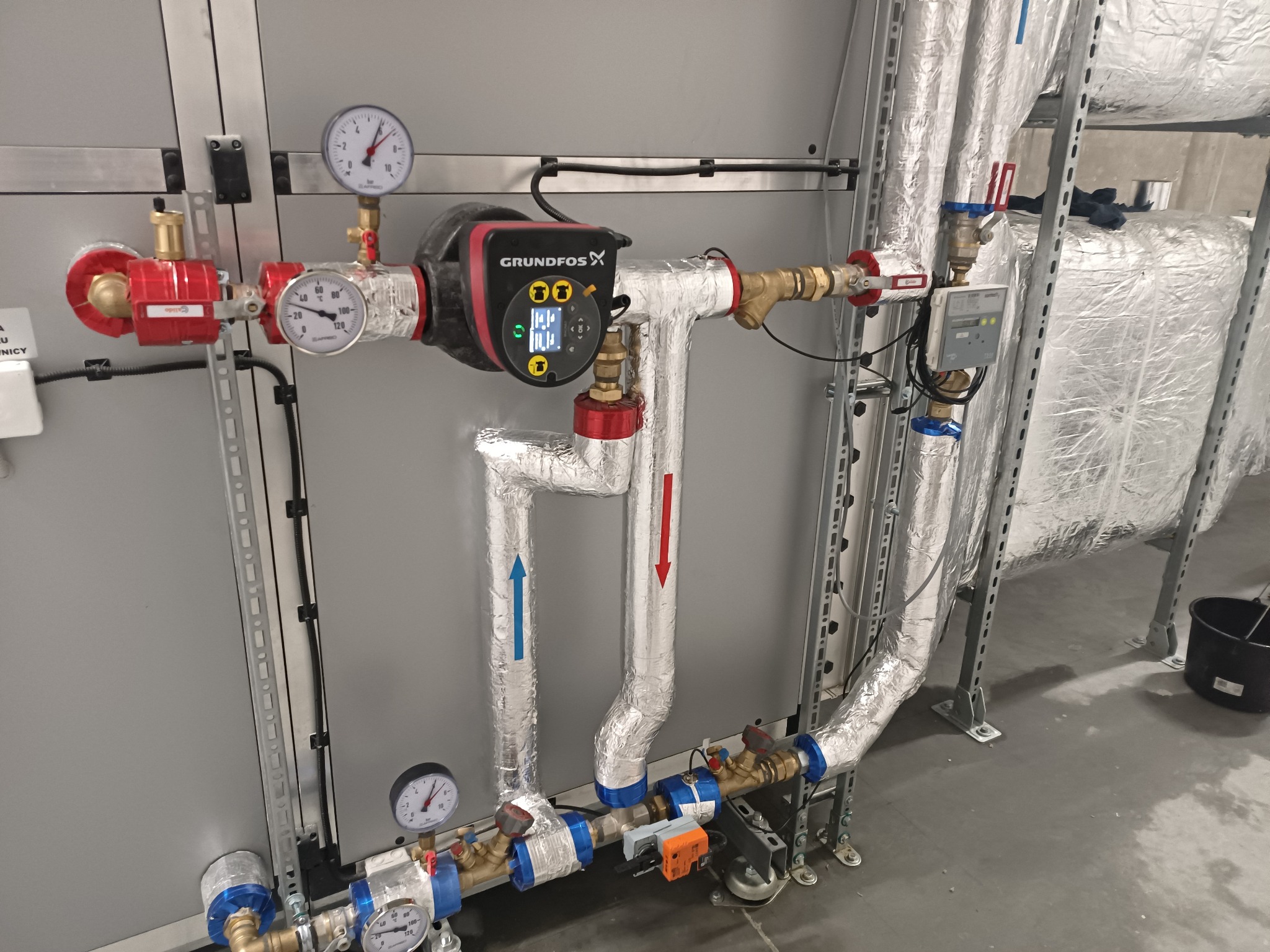 Węzeł cieplny z pompą obiegową Grundfos, manometrami i orurowaniem izolowanym folią aluminiową, widoczne strzałki wskazujące kierunek przepływu, zamontowany na szarej ścianie w pomieszczeniu...