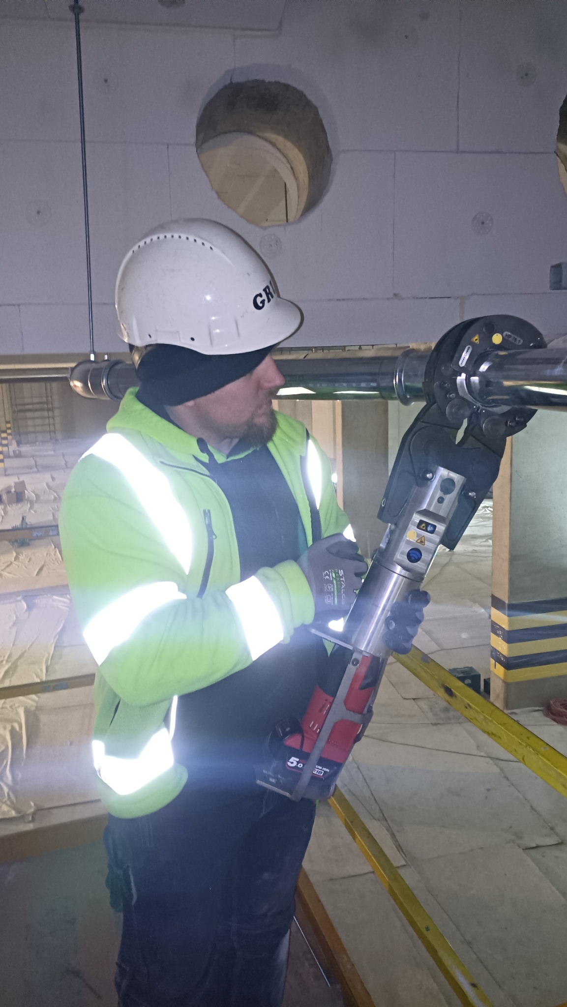 Pracownik w kasku i odblaskowej kurtce zaciska rurę hydrauliczną za pomocą specjalistycznej prasy akumulatorowej w surowym wnętrzu budynku z widocznymi otworami w ścianie.