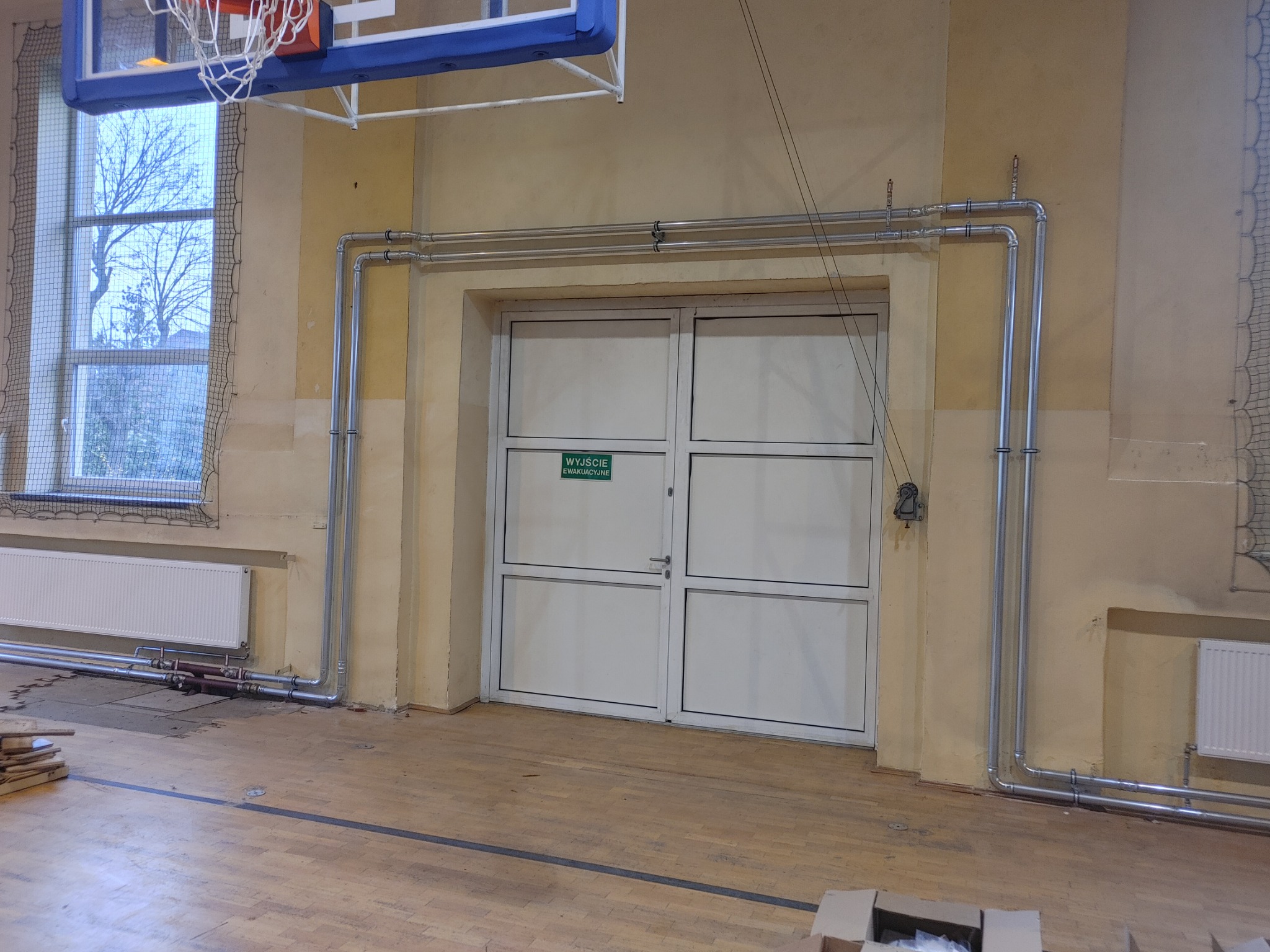 Instalacja grzewcza z rurami biegnącymi nad drzwiami ewakuacyjnymi w sali gimnastycznej, widoczne grzejniki i okno z siatką ochronną.