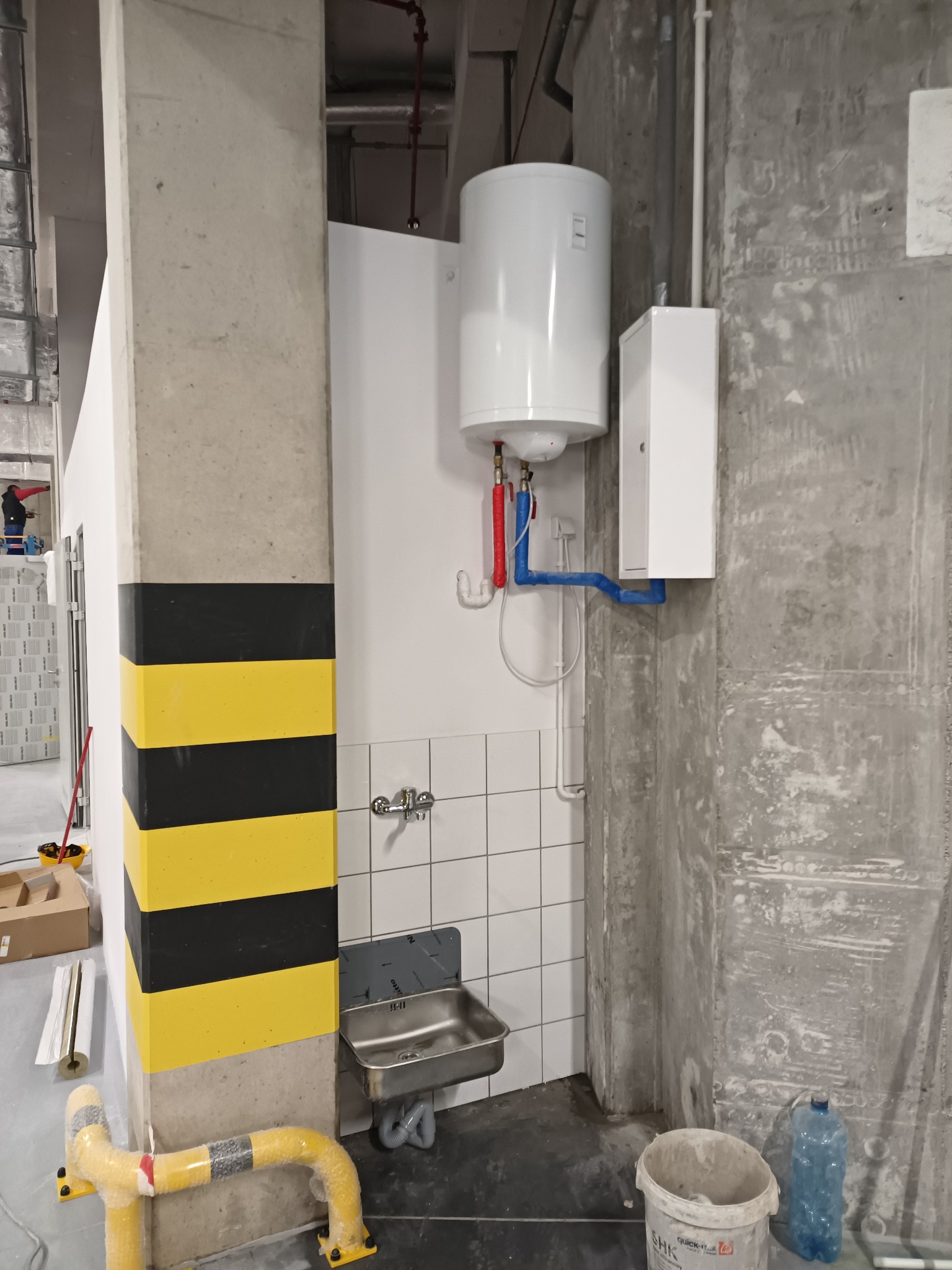 Bojler elektryczny zamontowany na ścianie z białą glazurą, widoczny zlew ze stali nierdzewnej i fragment instalacji wodnej z rurami w kolorze niebieskim i czerwonym, na pierwszym planie...