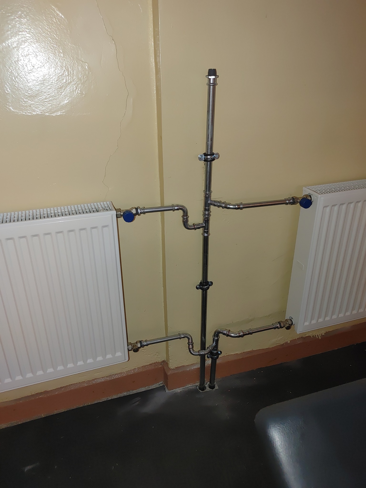 Instalacja grzewcza z rurami ze stali nierdzewnej podłączona do dwóch białych grzejników, widoczna na tle beżowej ściany z pęknięciem.